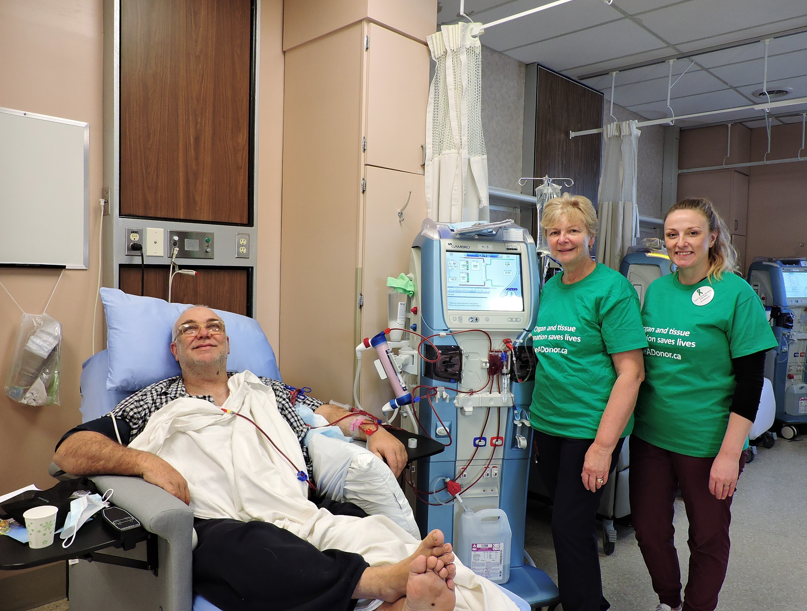Male patient receiving dialysis treatment in hospital with two female nurses in green organ donation shirts standing beside 